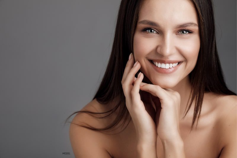 Brunette woman, smiling and touching her face against a gray backdrop.